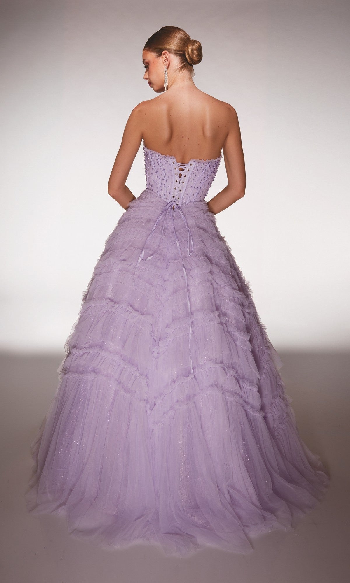 A woman stands facing away, her hair in a sleek bun, wearing the Alyce 62208 long prom dress with a strapless bodice, lilac tulle ruffle skirt, and lace-up back.